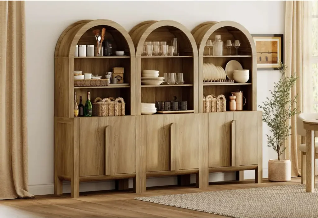 Frontal view of a three-piece light-oak arched cabinet unit, curated with varied ceramics and glassware, in a minimalist room with a potted olive tree.